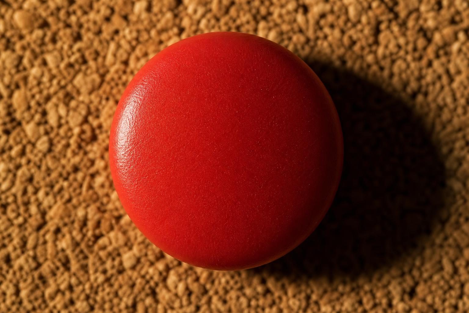 An extreme close-up shows a colored pushpin pressed into blank cork under hard light, with soft edges and a long shadow.