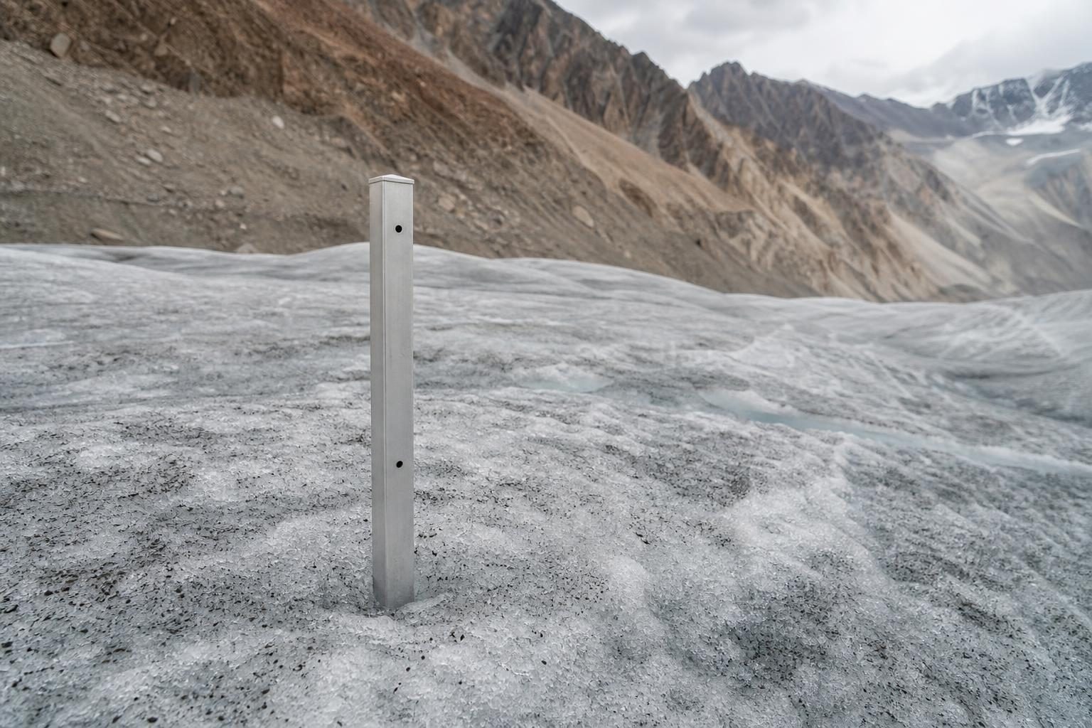 A measuring stake sticks out of thinning glacier ice below rocky mountain slopes.