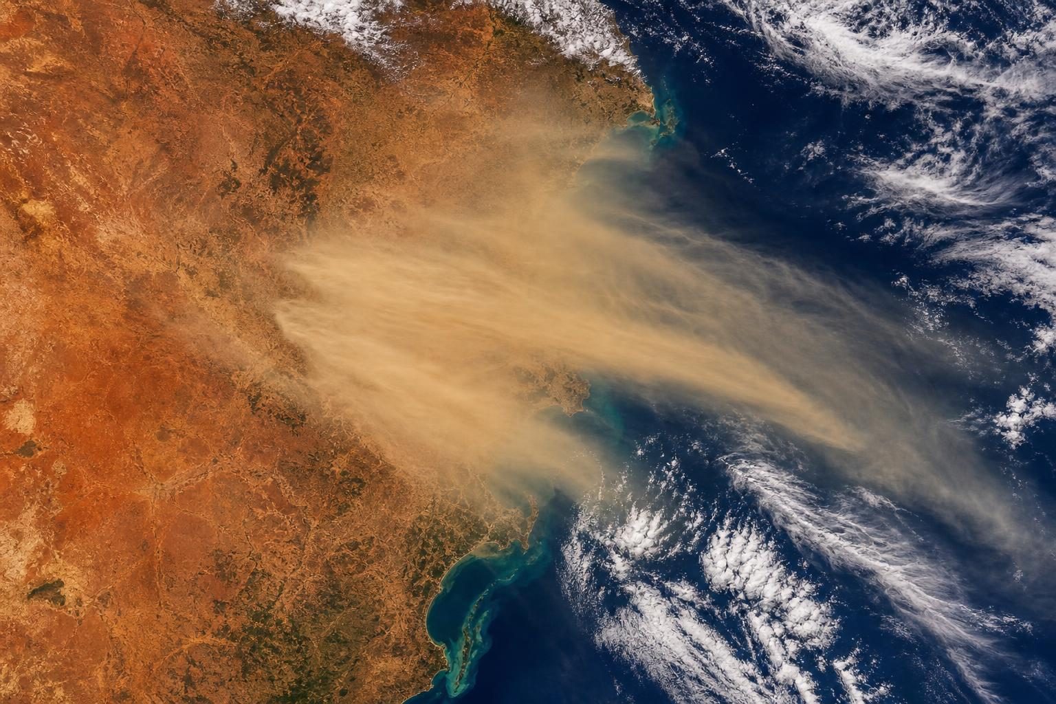 A broad tan dust cloud stretches over dry land toward the ocean as seen from above.